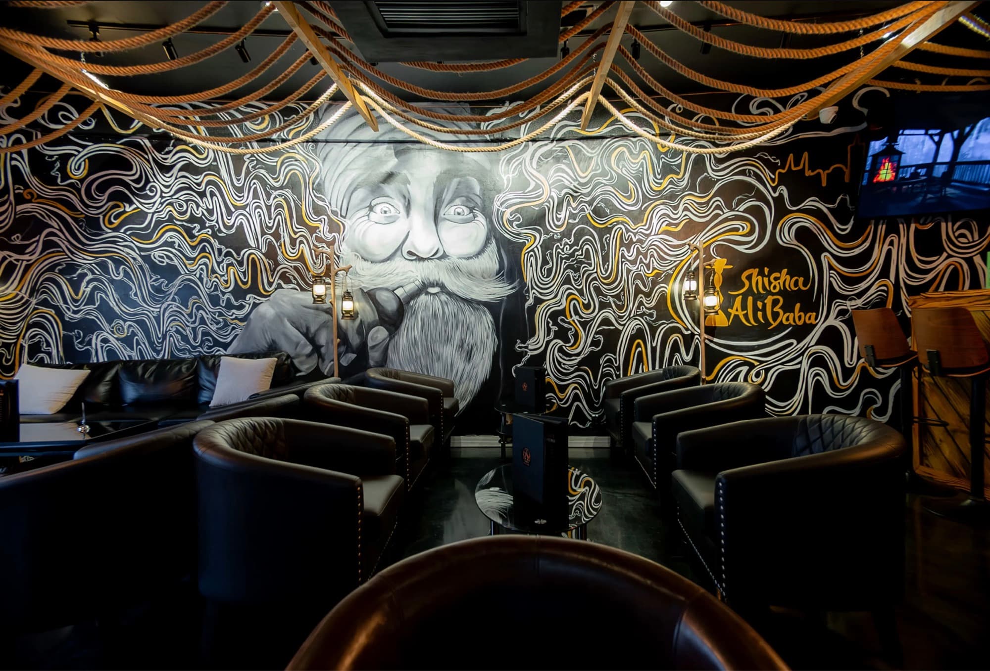 Mural wall of a bearded man with a pipe beside the Shisha Ali Baba logo, with leather lounge chairs in front
