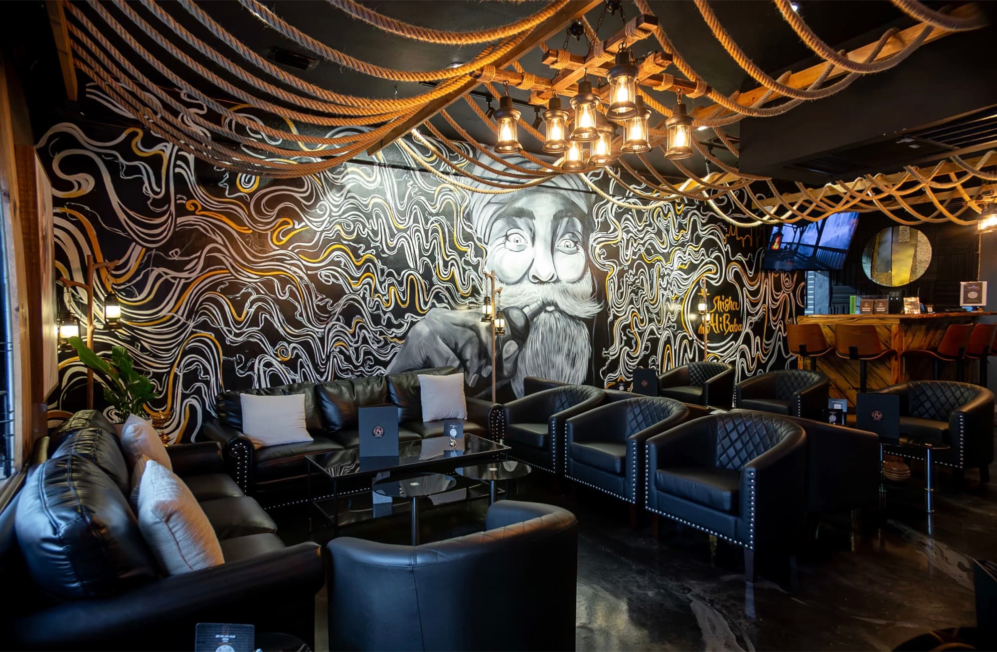 Black leather lounge chairs facing a black-and-white mural under rope and lantern ceiling lights