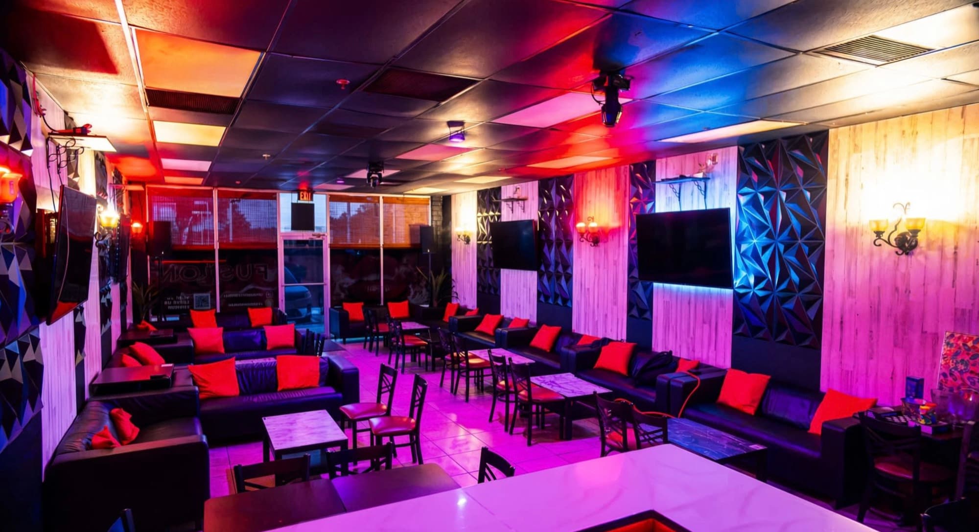 Wide view of the lounge with leather sofas, orange pillows, marble tables and wall TVs under neon lighting